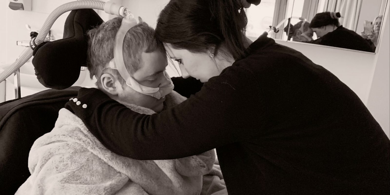Mum and son embracing with heads rested together and eyes closed, son is in a wheelchair wearing breathing apparatus, wrapped up in a blanket and mum is wearing black cardigan with hair in a bun