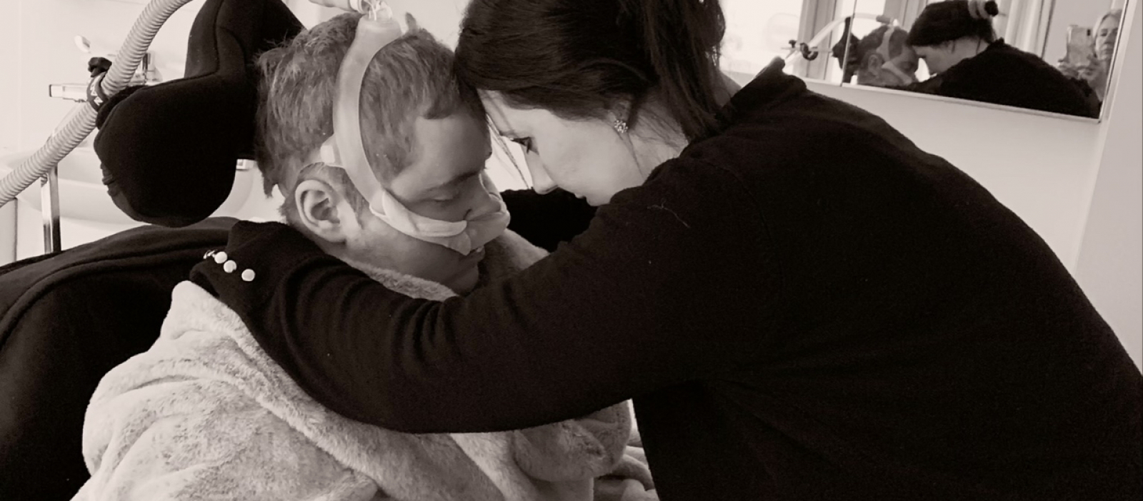 Mum and son embracing with heads rested together and eyes closed, son is in a wheelchair wearing breathing apparatus, wrapped up in a blanket and mum is wearing black cardigan with hair in a bun