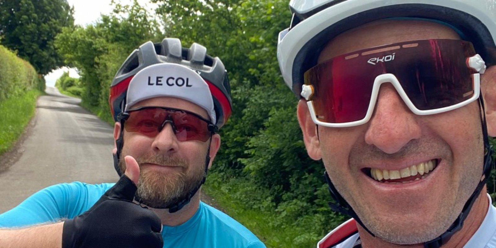 Lee and Sylvain, two men, taking selfie wearing helmets, sunglasses and cycling attire, in front of green trees with thumbs up